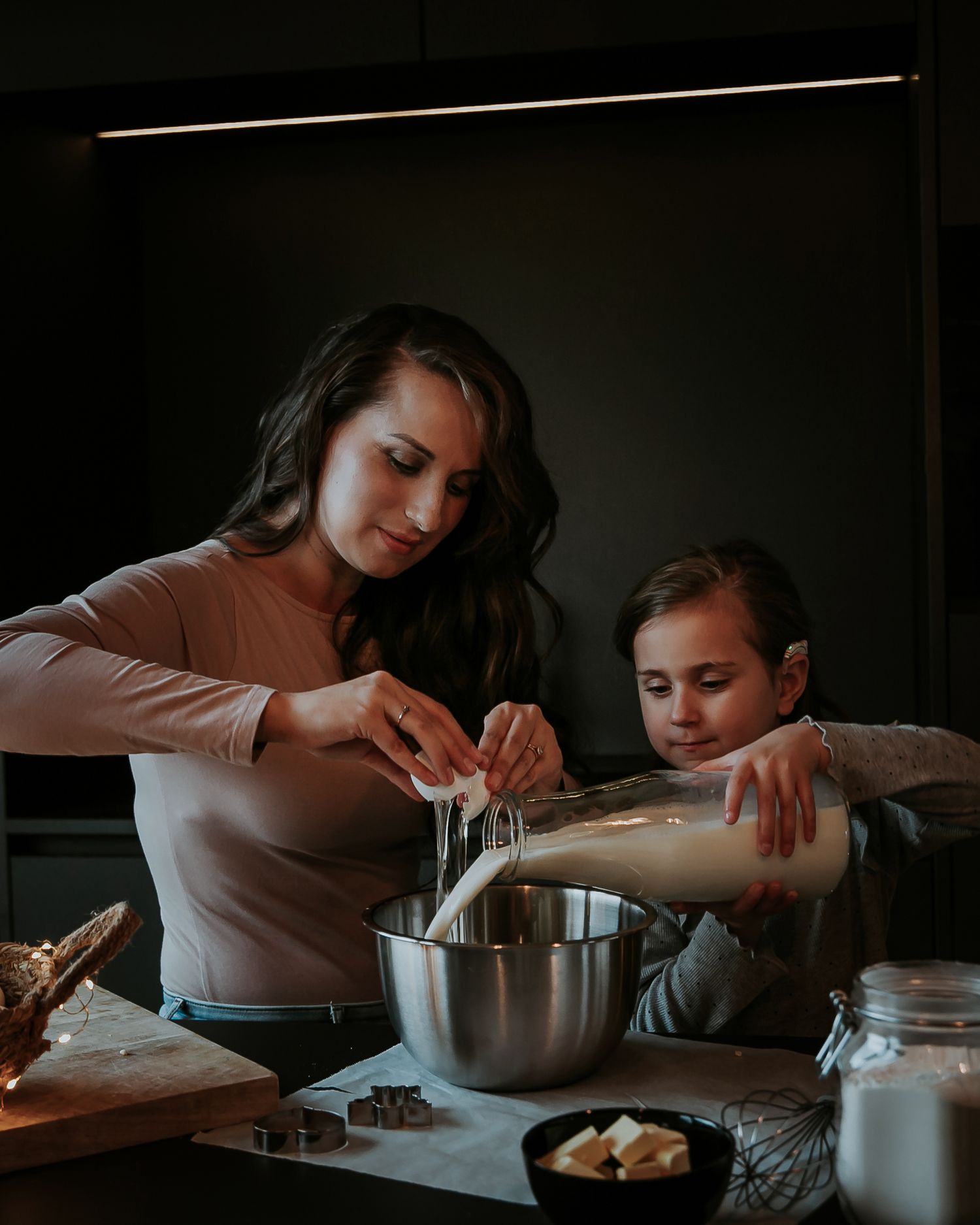 Mother and daughter bake holiday cookies together at a cozy kitchen table with milk, flour and with twinkling lights behind them in Woerden.