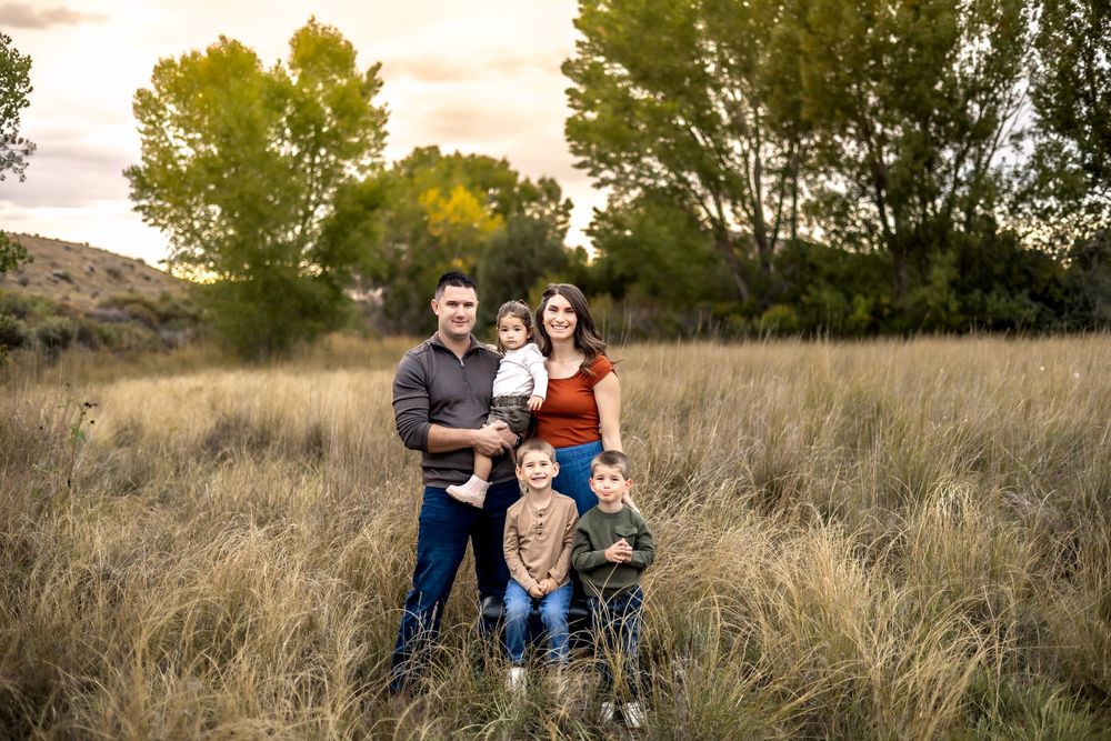 A smiling couple holding a toddler stands with two young boys in a field of tall autumn grass under green trees.