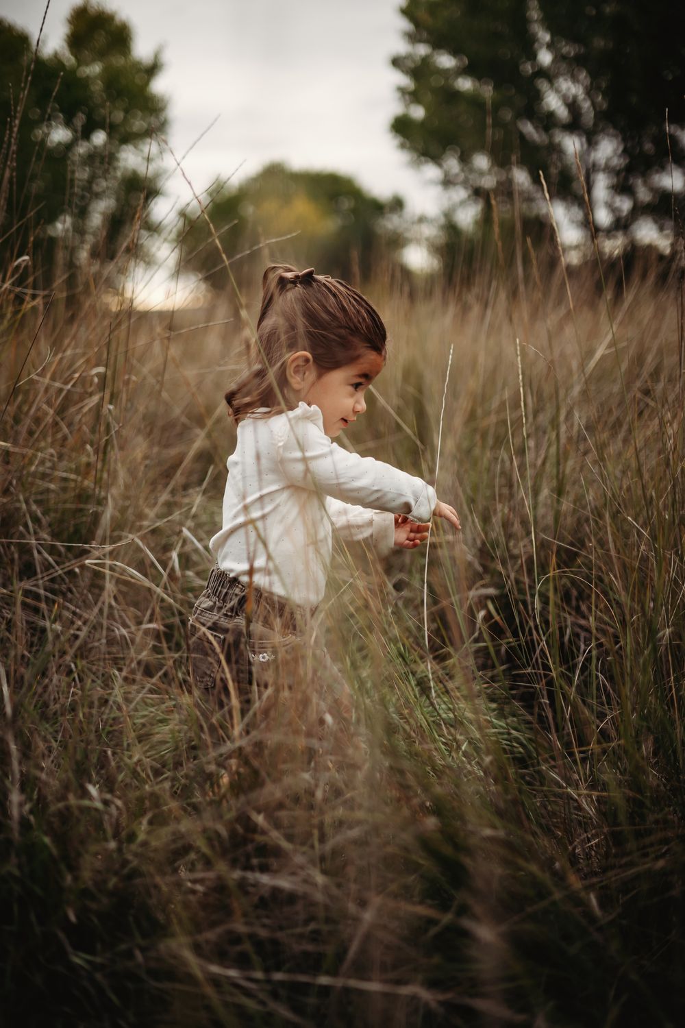 Young girl in a cream blouse with a small ponytail walks through tall golden grass in a sunlit field.