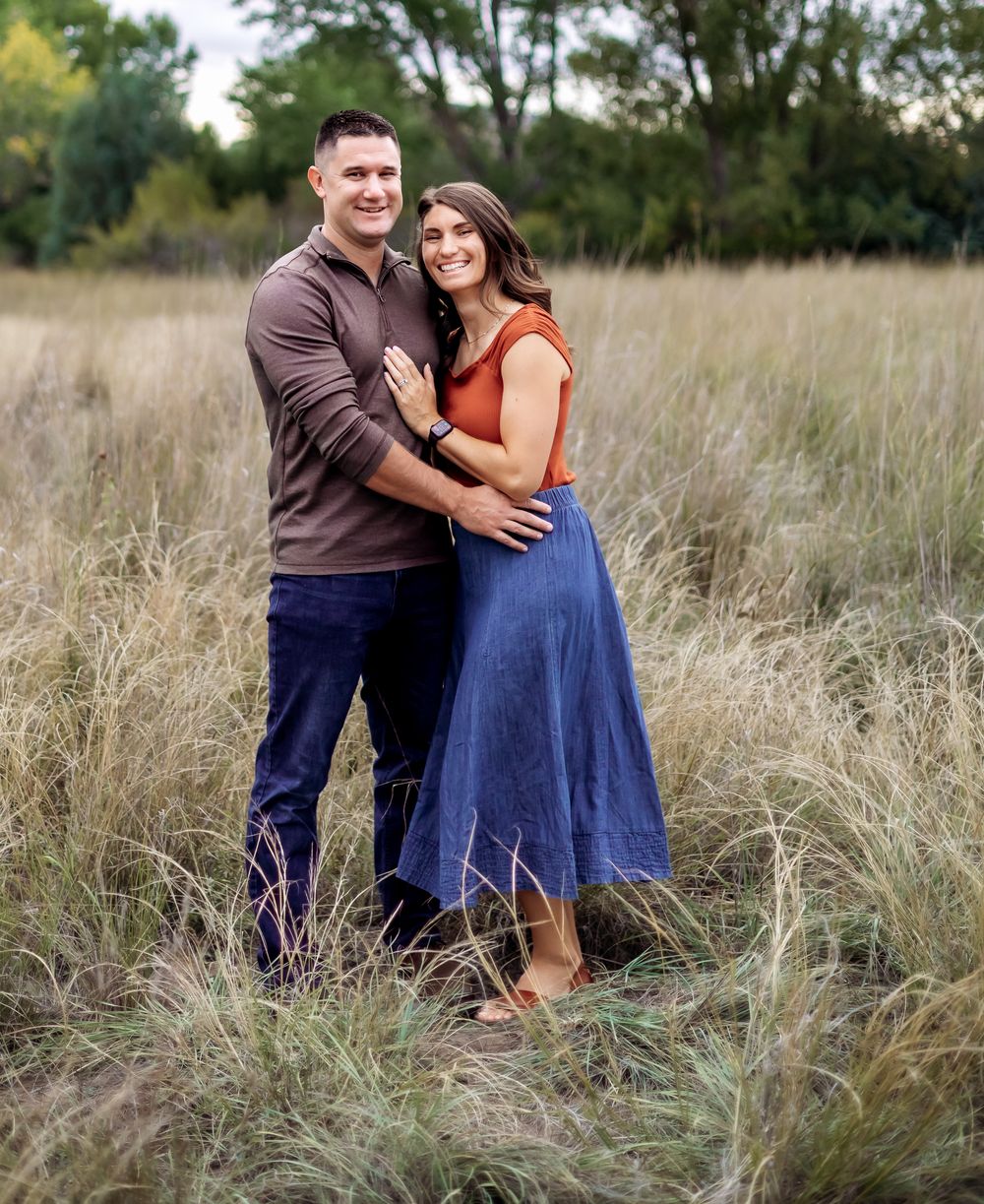 Smiling couple embraces in a tall grass field, he in a brown shirt and jeans, she in an orange top and blue skirt.