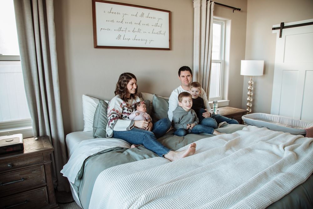 A family lifestyle photo shows parents, two toddler boys and a baby sitting together on a bed in a bright bedroom.