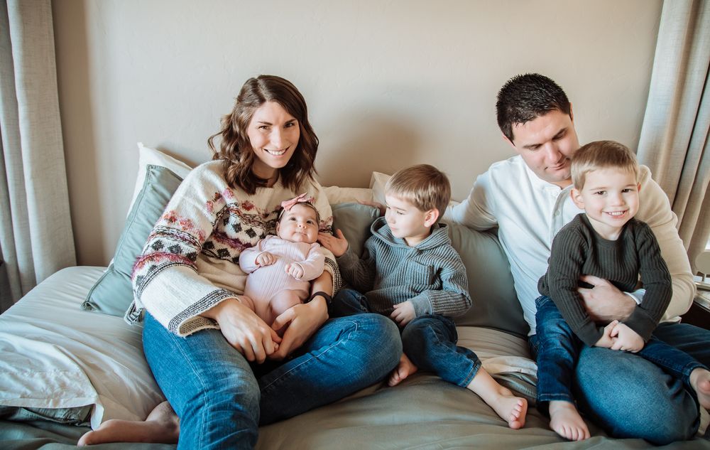 A happy family of five snuggles on a bed as the mother holds her baby girl while the father and two sons smile.