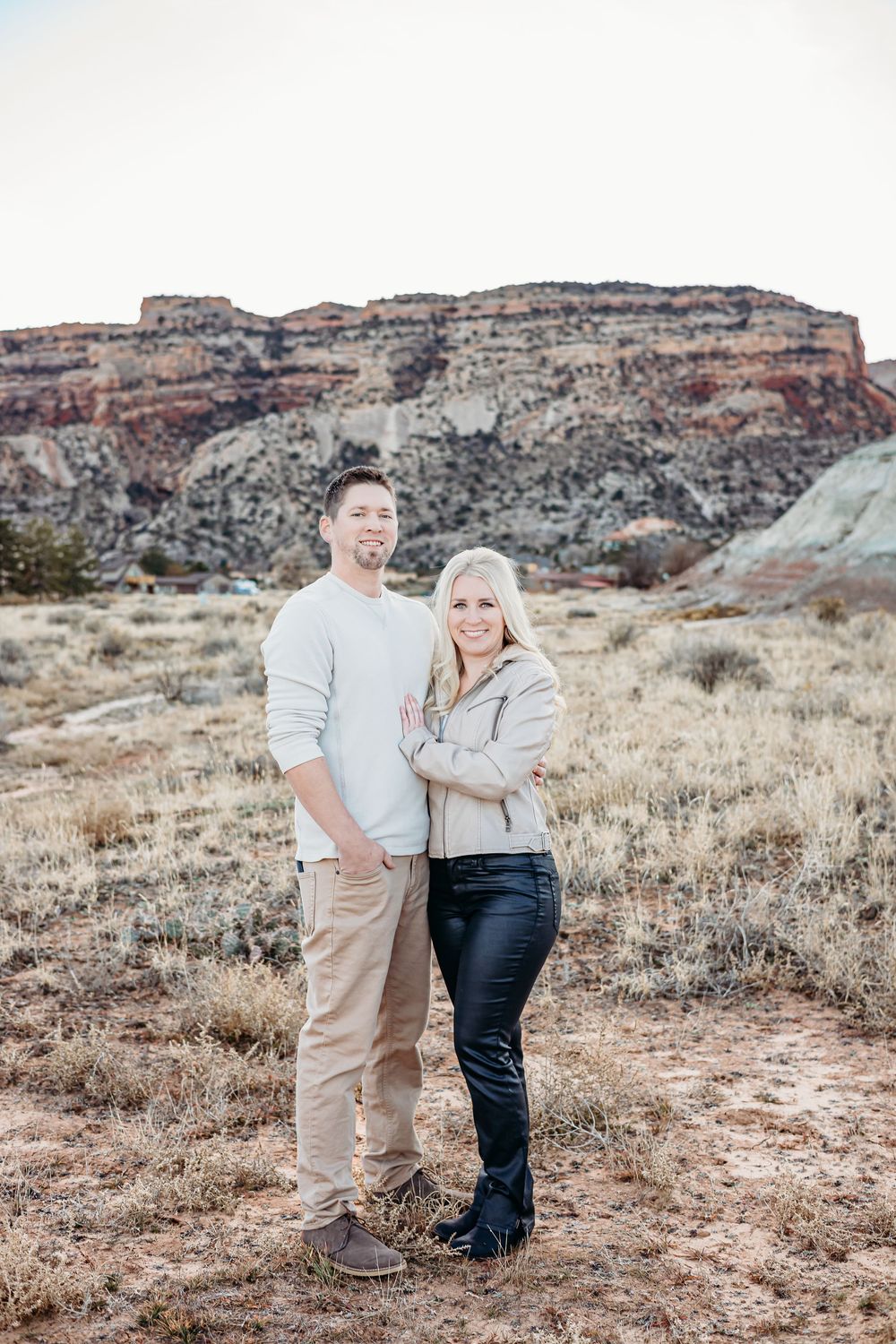Smiling couple embracing outdoors in front of red rock cliffs and dry desert grass during an engagement or family photo session.