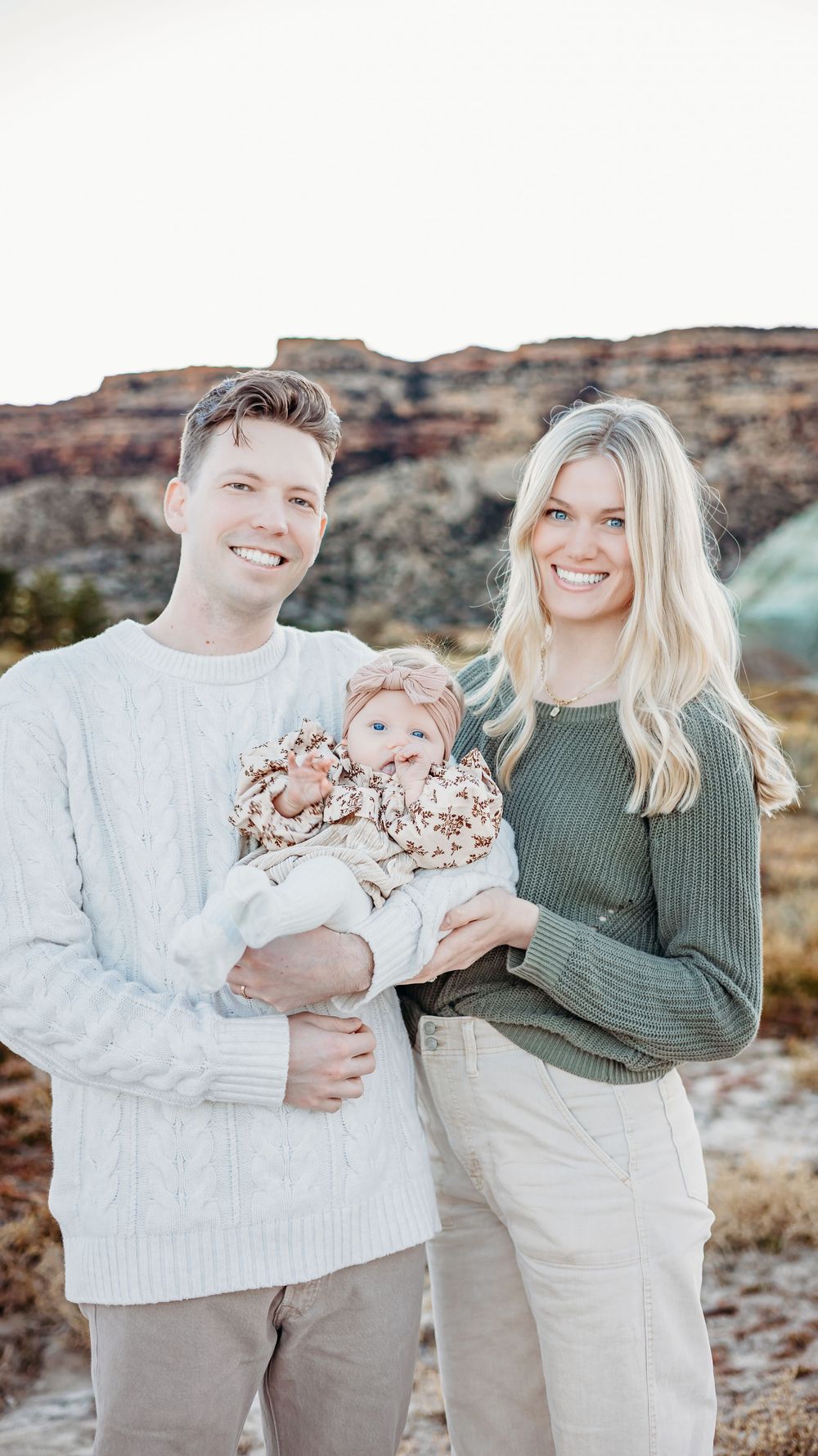 Smiling couple in cozy sweaters hold their baby girl in a floral outfit with red rock cliffs behind them in the desert.