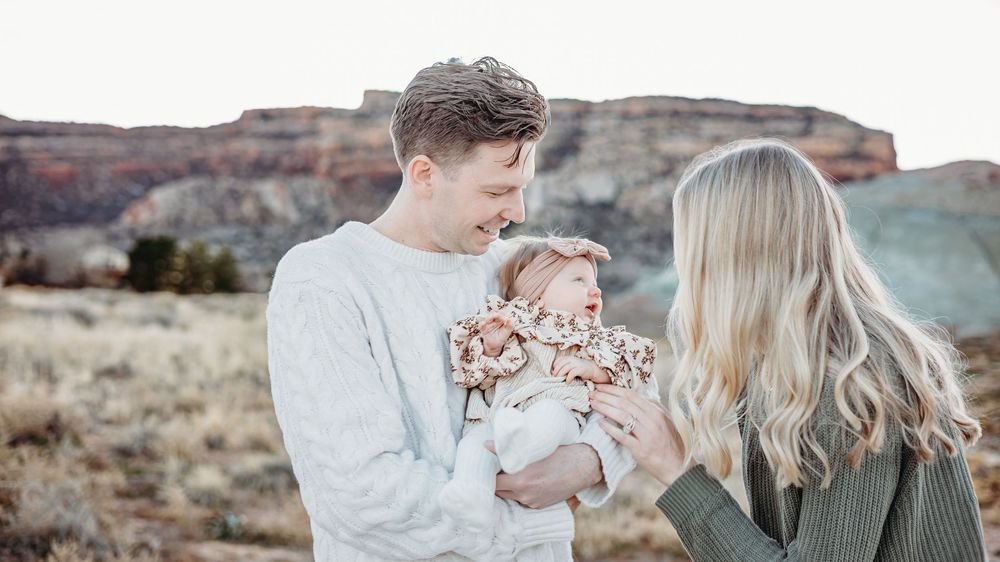 Father holds his baby girl in a floral outfit while the mother with long blonde hair looks on, with red rock cliffs behind.