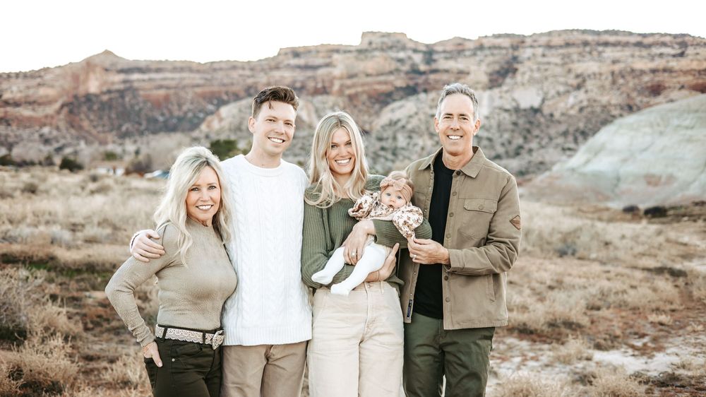 Multigenerational family of five with a baby, smiling together in front of red rock cliffs in a desert landscape.