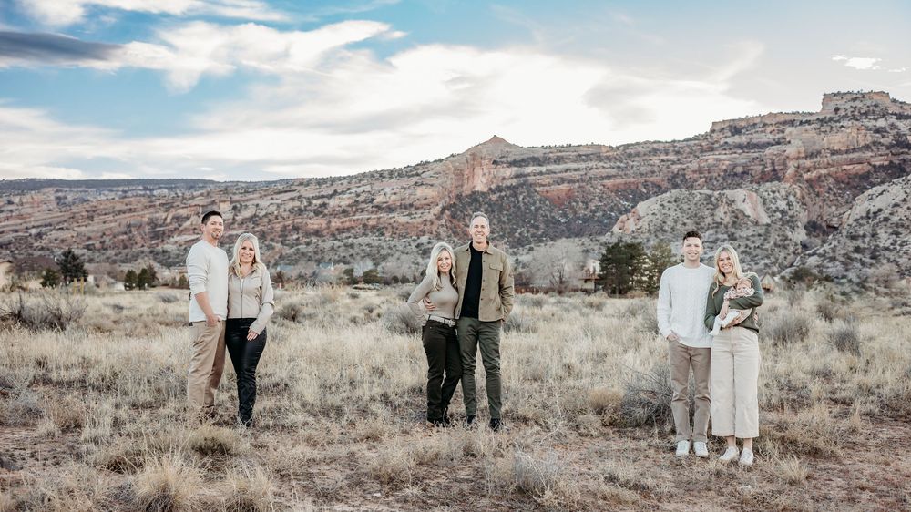 A family stands in three small groups across a wide desert landscape with red rock cliffs and a cloudy sky behind them.