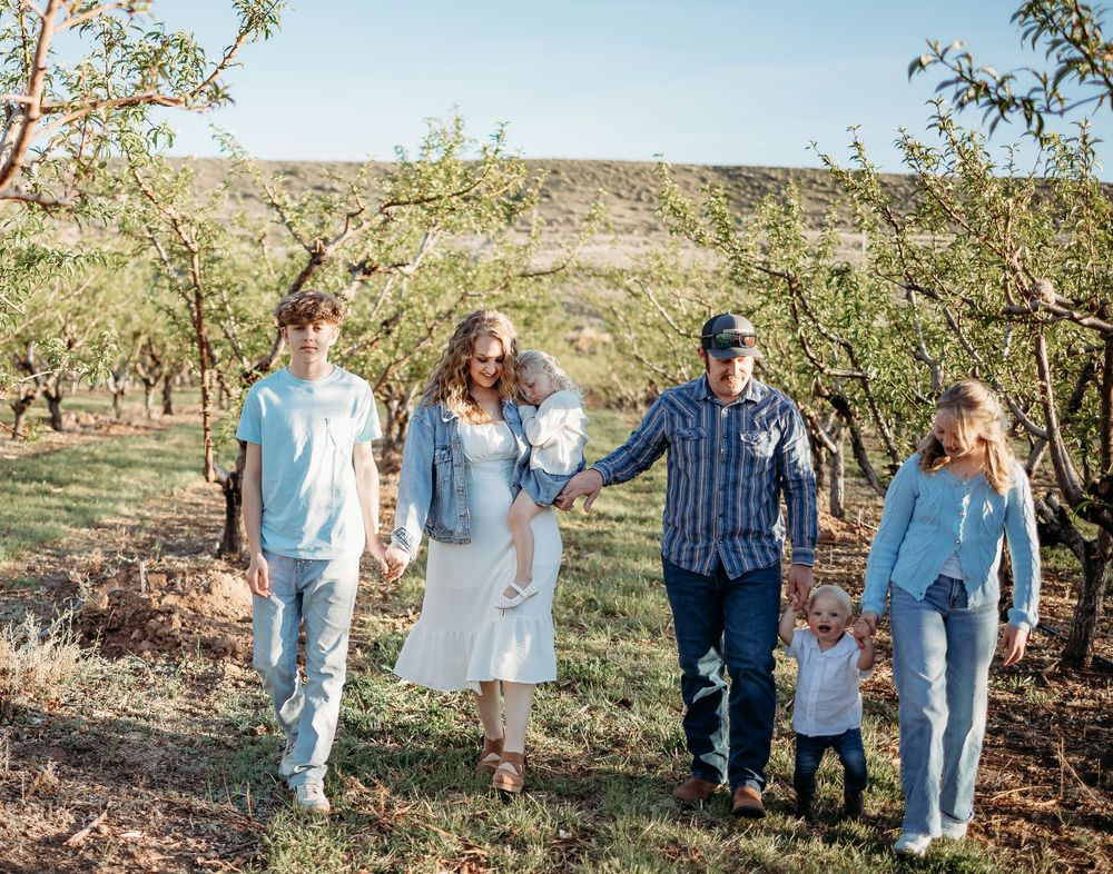 A family walks hand in hand through an orchard of fruit trees, with parents carrying a baby and holding a toddler beside two teens.