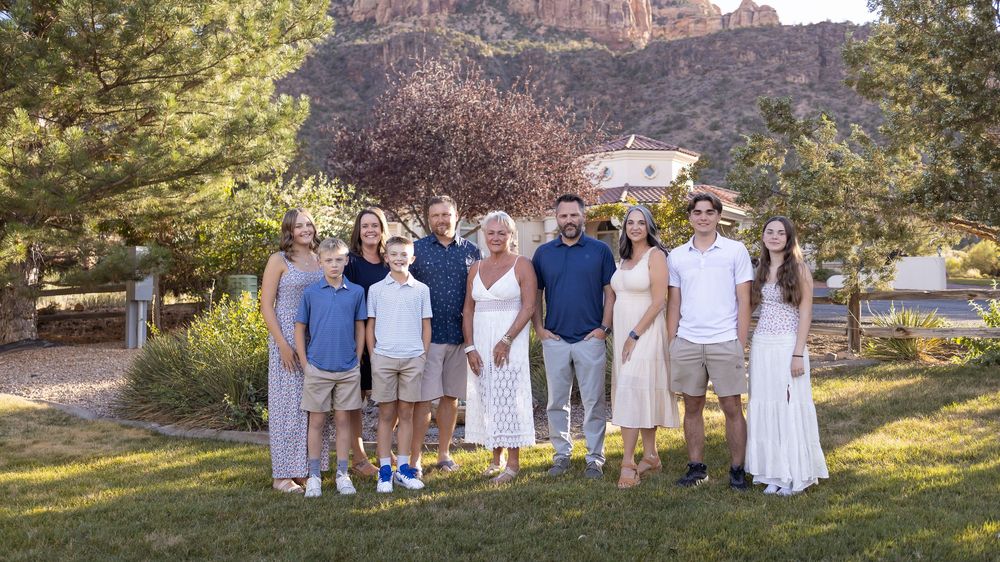 A large extended family of eleven poses together on a grassy lawn in front of towering red rock cliffs and autumn trees.