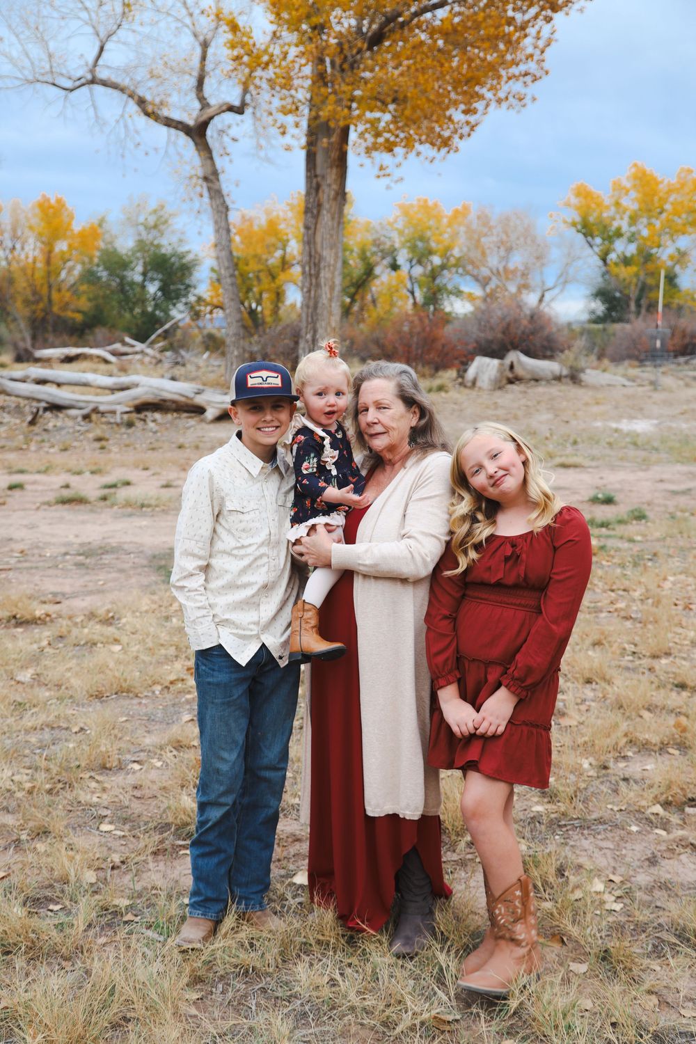 Grandmother and three grandchildren in cowboy boots stand together under a golden autumn tree for a family portrait.