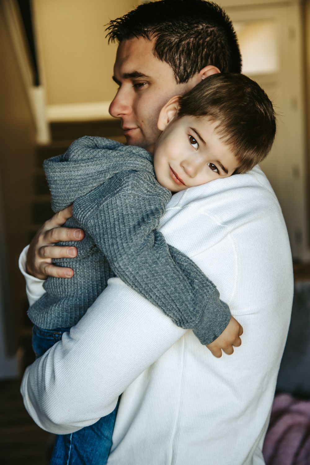 A father in a white sweater embraces his sleepy young son in a gray knit sweater, resting his head on dad's shoulder at home.