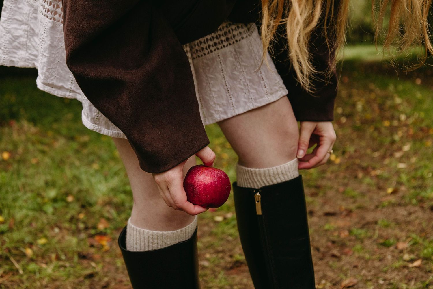 Woman in a white skirt, brown jacket and black knee-high boots with knit cuffs holds a red apple in an autumn park.