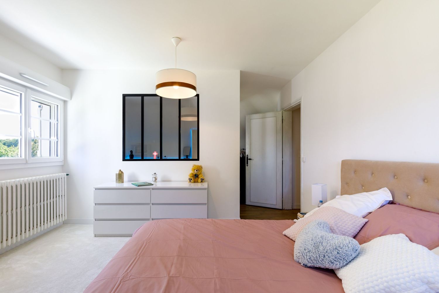 Chambre lumineuse avec une literie rose, une commode blanche, une suspension et une fenêtre intérieure donnant sur la salle de bain