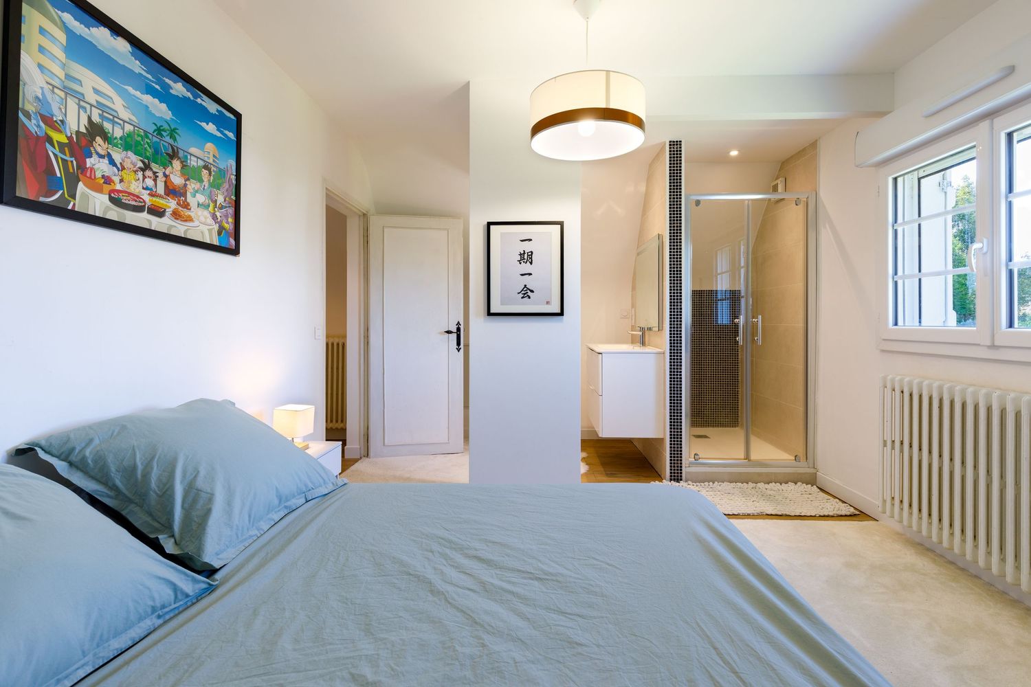Chambre avec literie grise, salle de douche attenante, décorations murales et lumière naturelle provenant d'une fenêtre.