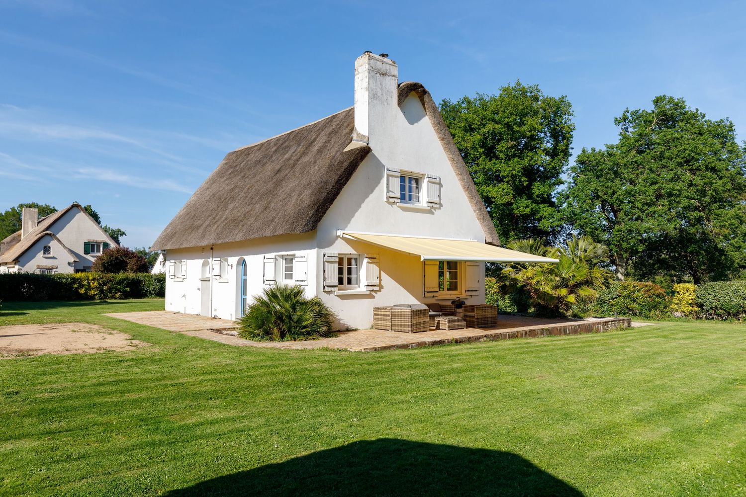 Charmant cottage au toit de chaume et aux murs blancs, doté d'une pelouse verdoyante et d'une voile d'ombrage jaune au-dessus de la terrasse