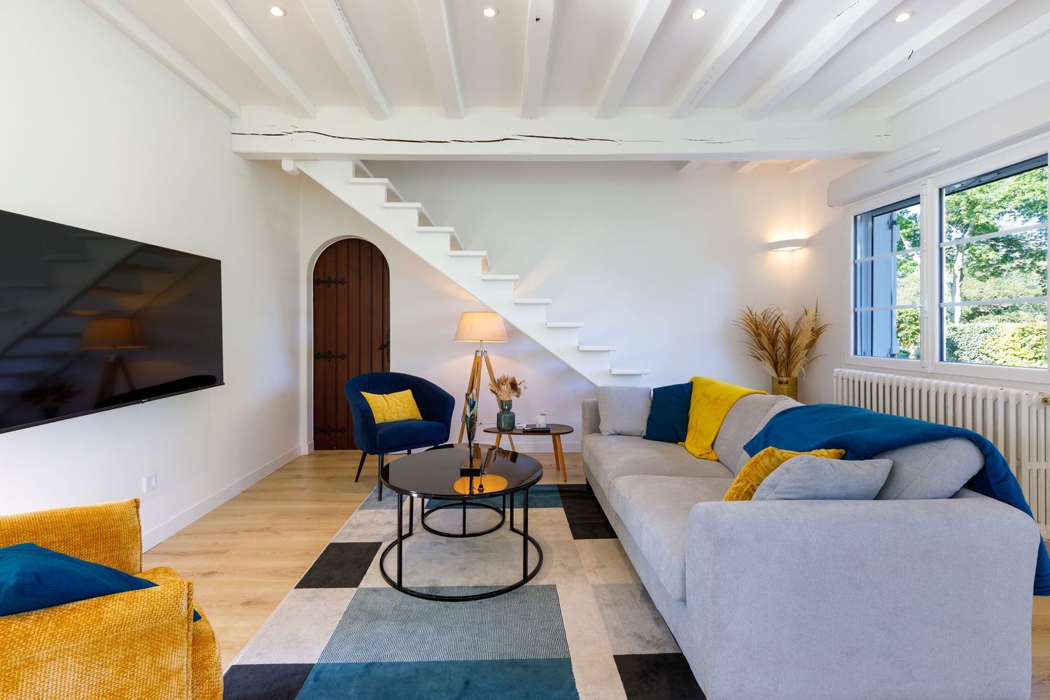 Salon avec canapé gris, touches de jaune, tapis géométrique, escalier et grande fenêtre laissant entrer la lumière naturelle.
