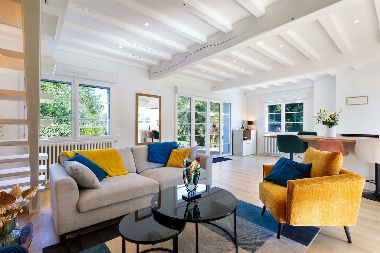 Séjour lumineux avec plafond à poutres apparentes, canapé gris, fauteuils en velours doré, coussins bleus et tables basses en ver