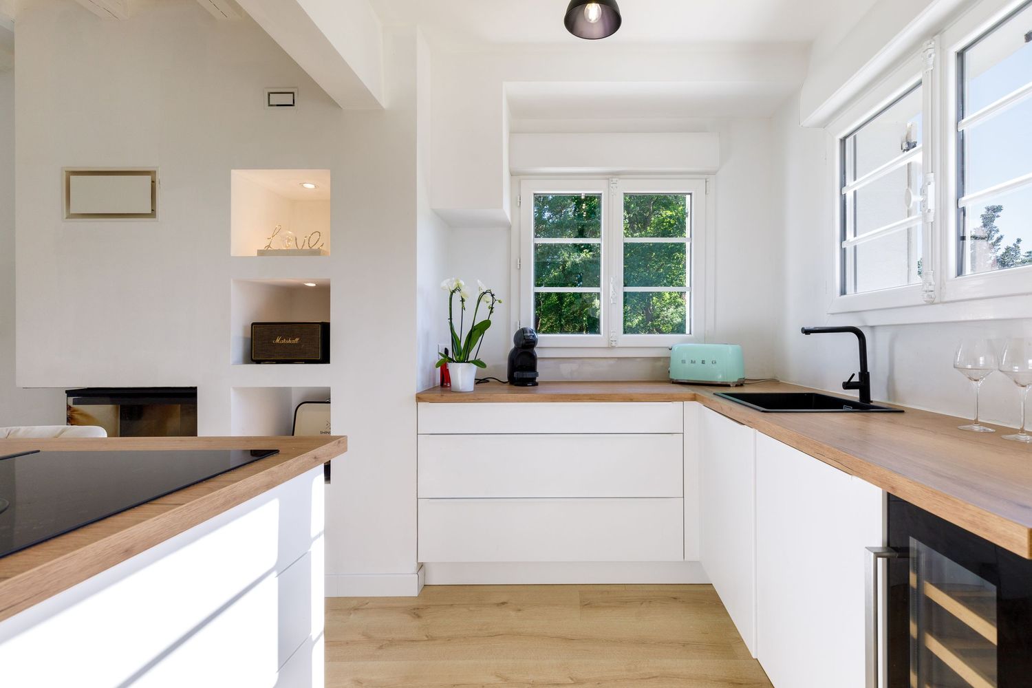 Cuisine blanche avec plans de travail en bois, robinetterie noire, cave à vin et une fenêtre lumineuse donnant sur de la verdure.
