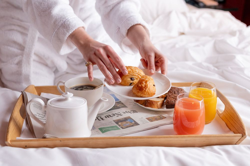 Mains en peignoir blanc posant une assiette de viennoiseries sur un plateau en bois avec café, théière et jus, au lit