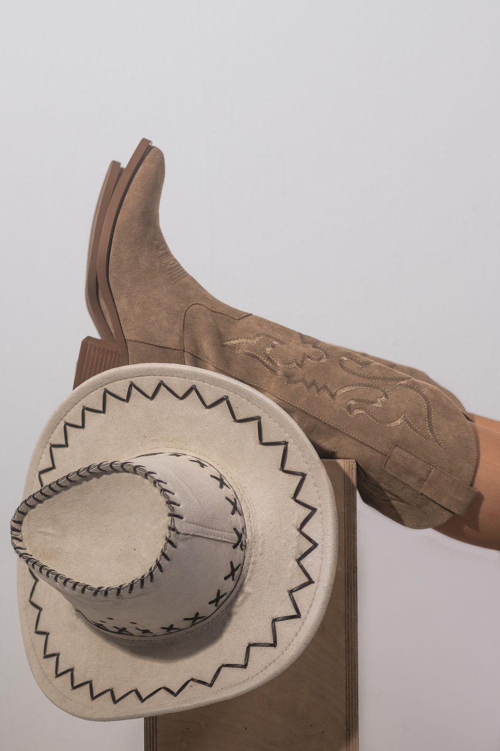Bottes de cowboy en daim beige à broderies, jambes levées, posées sur un chapeau western crème à coutures noires