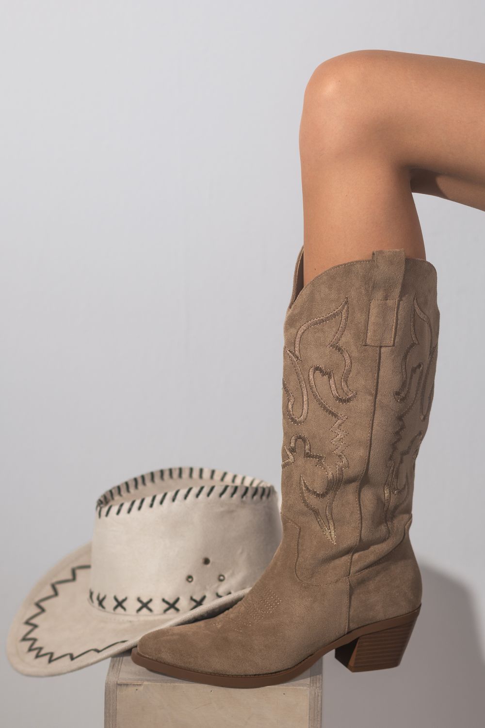 Jambe croisée dans une botte de cowboy en daim beige brodée, posée sur un cube en bois près d'un chapeau crème