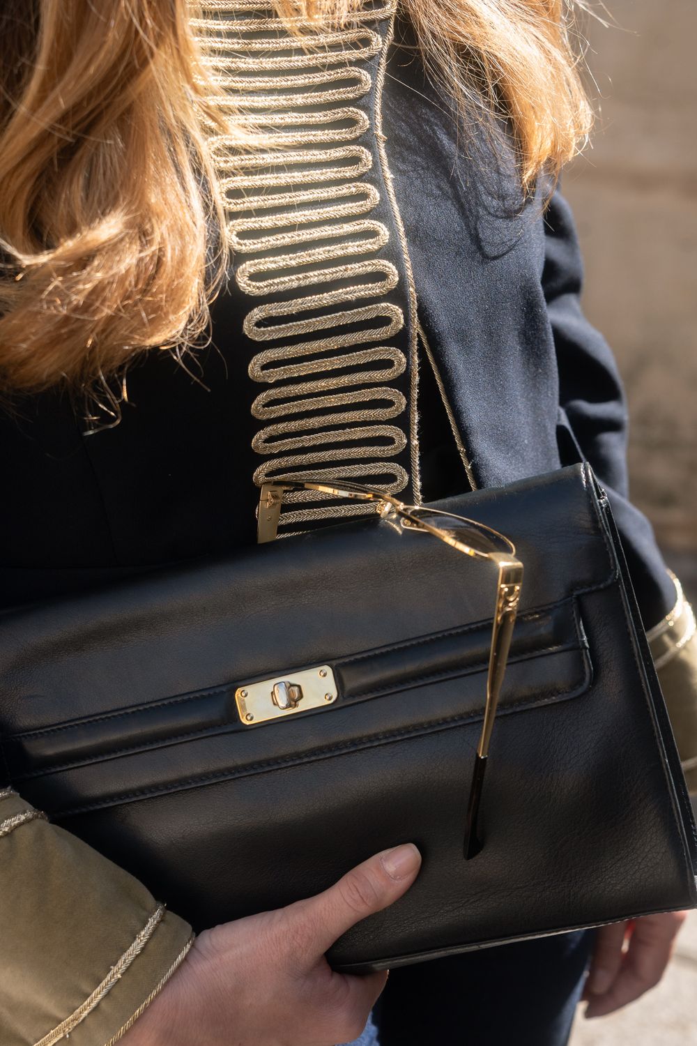 Femme aux cheveux blond vénitien, veste marine à galons dorés, pochette en cuir noir et lunettes dorées en main