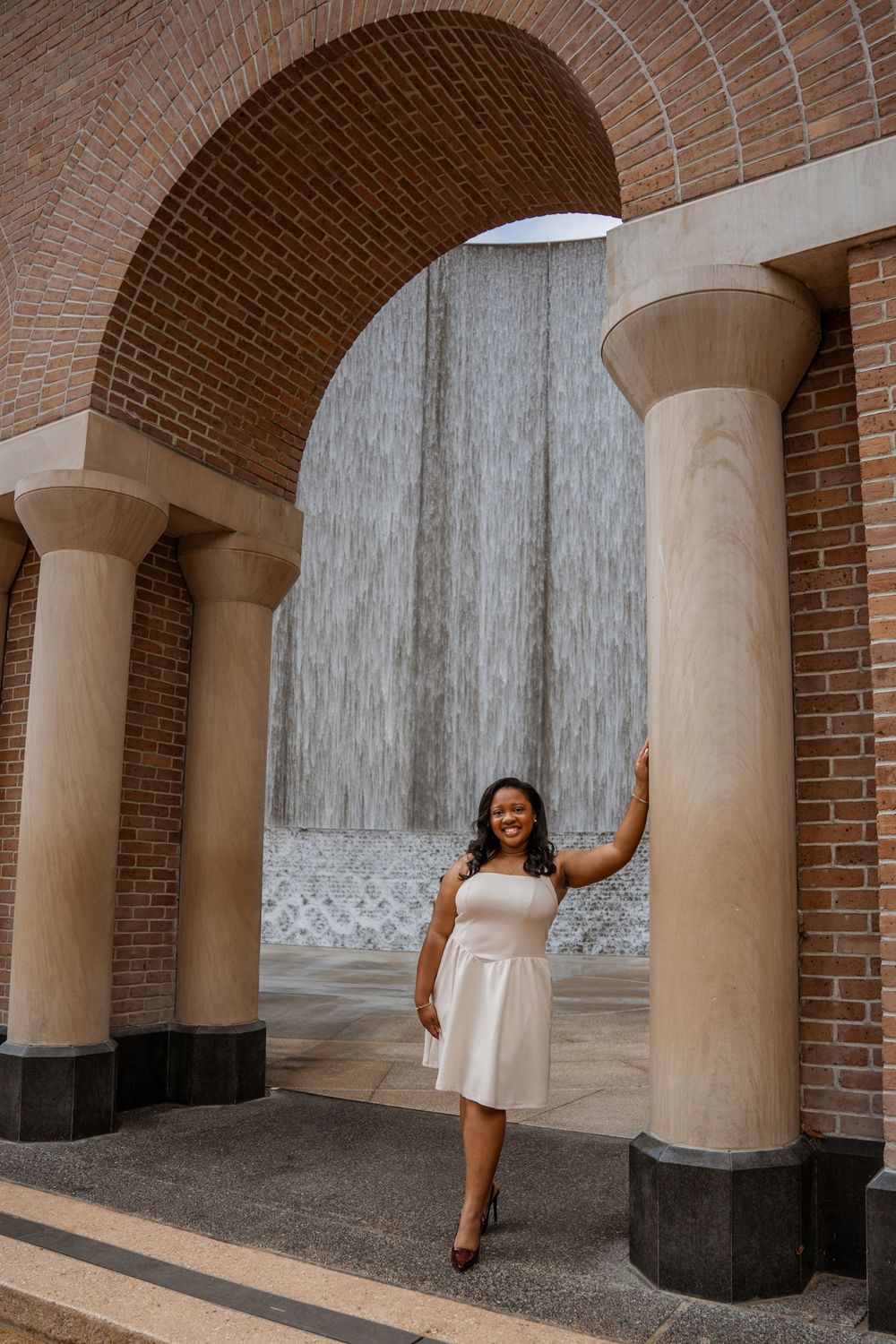 Young woman in white dress posing under brick arch with waterfall backdrop.