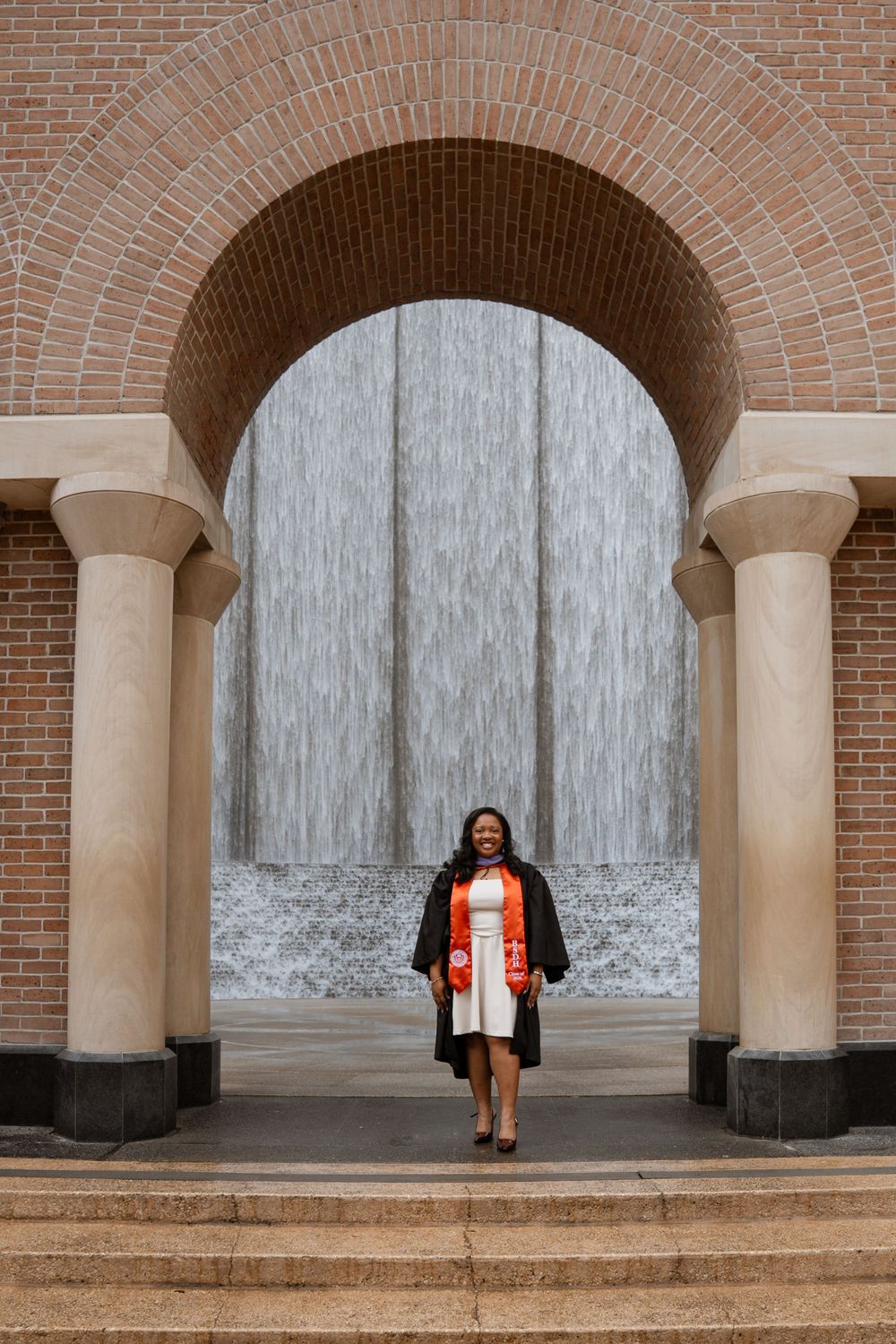 Graduate in black gown and orange sash smiles under brick arch with waterfall backdrop on graduation day.