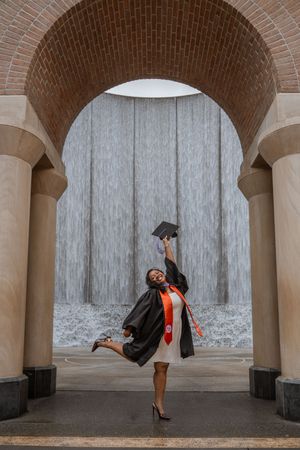 Graduate in cap and gown celebrates under brick arch with waterfall backdrop, joyfully raising mortarboard.