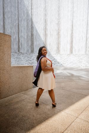 Smiling woman in white dress with purple bag poses in front of a waterfall wall on a sunny day.