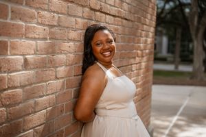 Smiling young Black woman in a cream dress leaning against a brick wall outdoors.