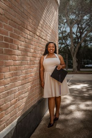 Smiling graduate in beige dress holding cap against brick wall outdoors.