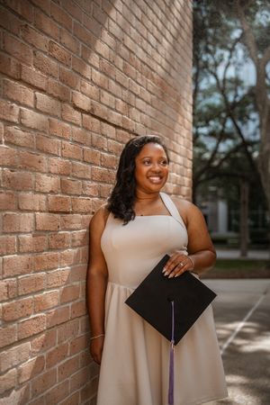 Smiling graduate in beige dress holding black cap against brick wall outdoors.