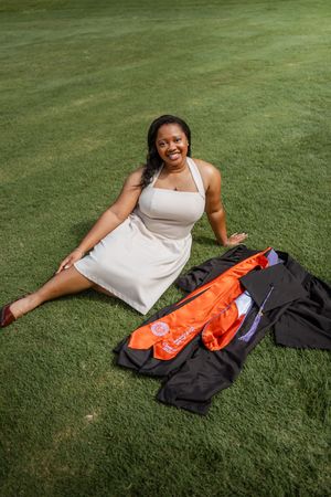 Smiling Black woman in white dress sitting on grass with graduation gown and orange sash beside her.