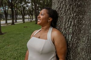 A smiling Black woman in a white sleeveless top leans against a tree trunk in a lush green park.