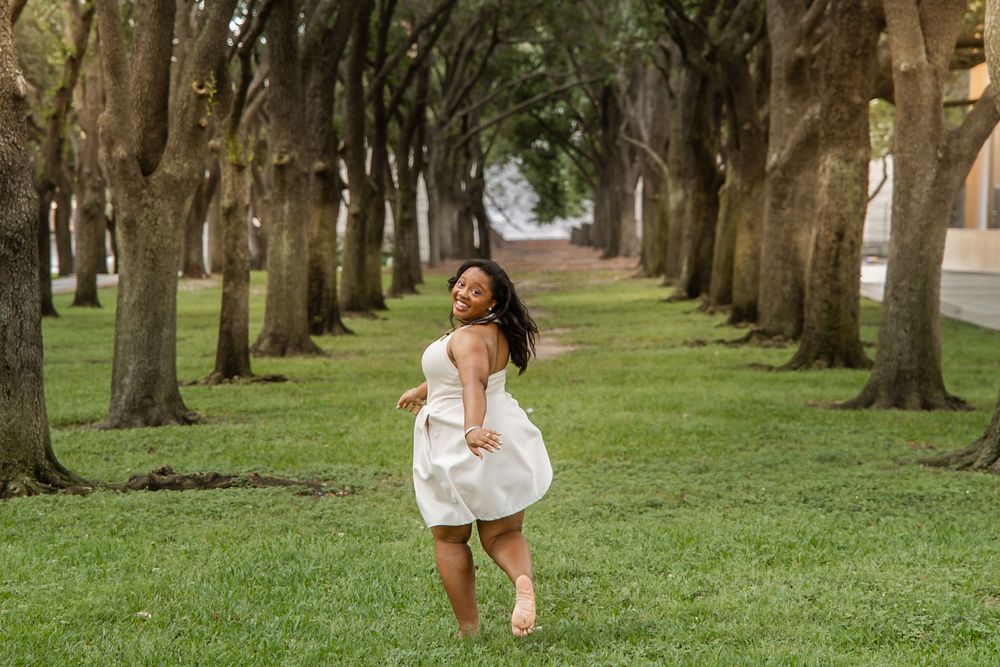Young Black woman in white dress twirling joyfully among tree-lined path on green lawn.