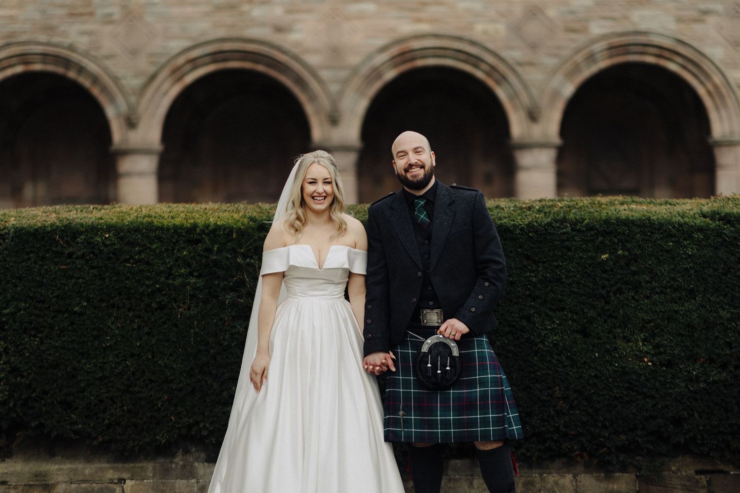 Smiling bride and groom in tartan kilt posing before stone arches at a Scottish wedding.