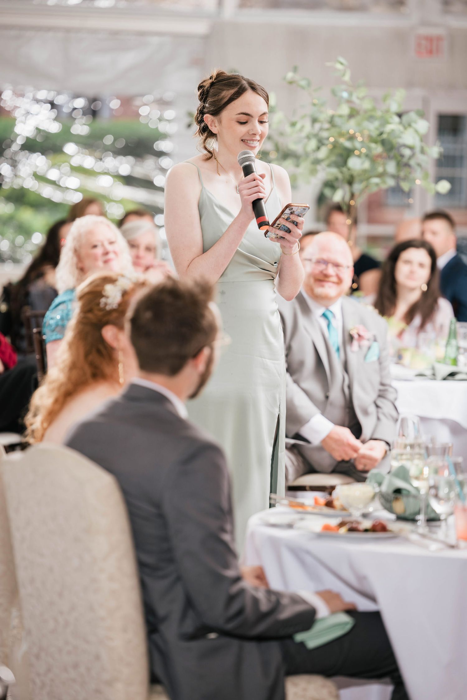 Bridesmaid giving a speech from her phone, guests seated around her smiling.