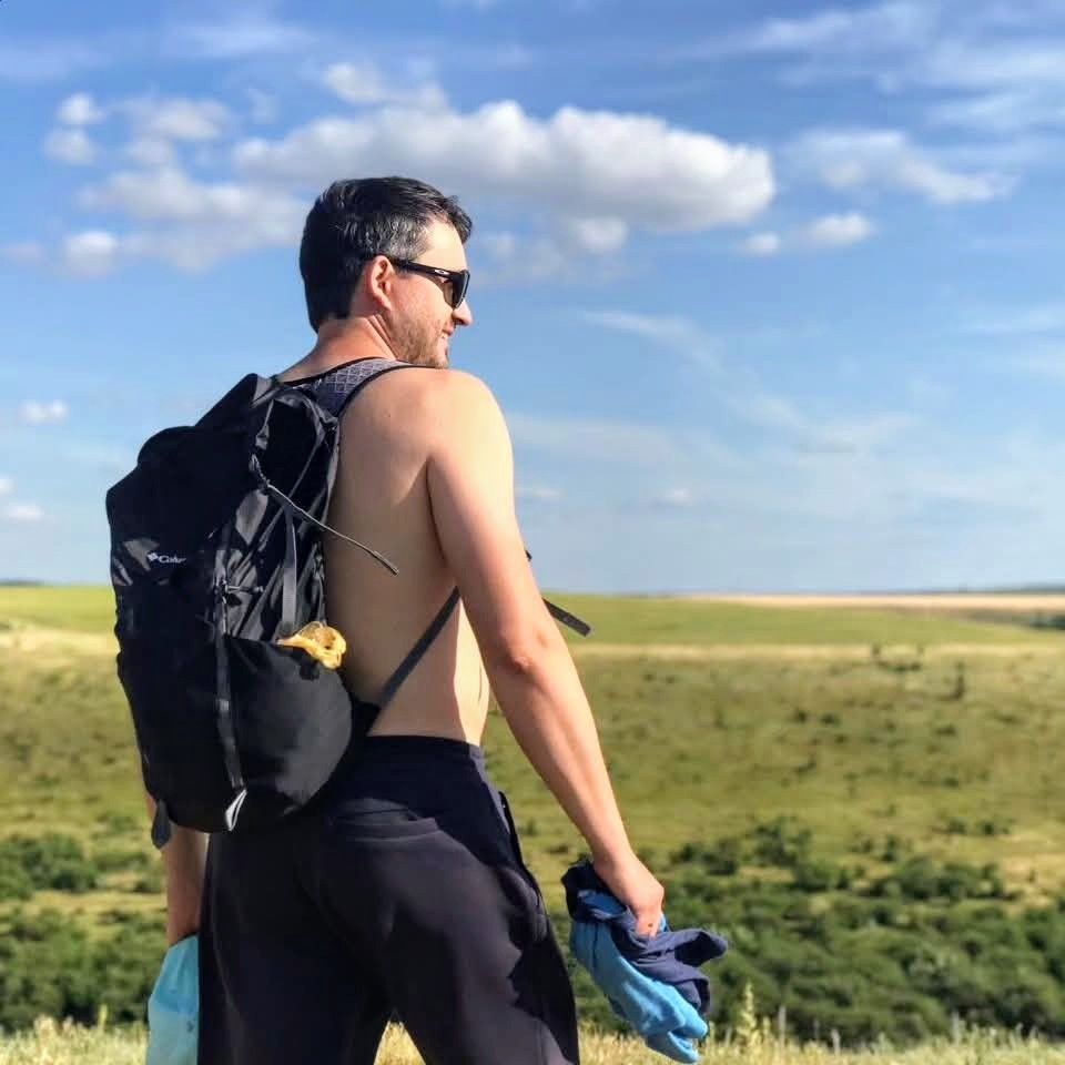 Shirtless male hiker with backpack and sunglasses enjoying panoramic views of green rolling hills.