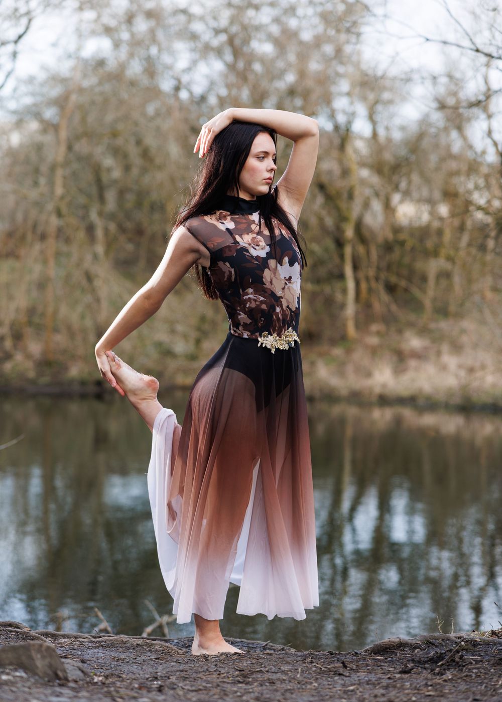 Barefoot dancer in floral lace bodysuit and ombre skirt poses gracefully outdoors near a reflective pond.
