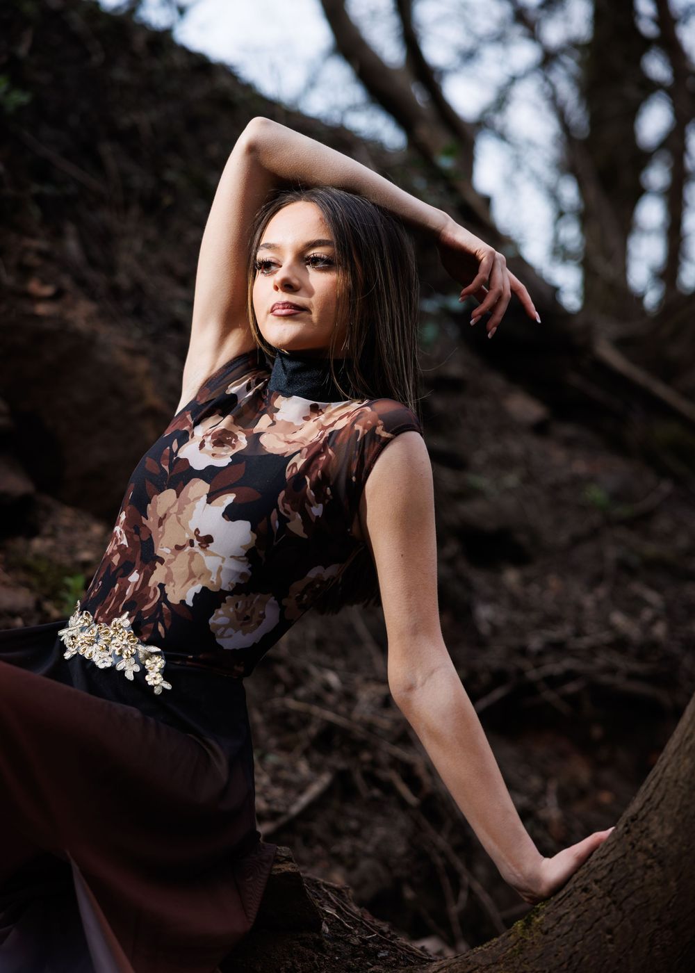 Young woman in floral dark dress poses gracefully among trees in a forest, arm raised elegantly.