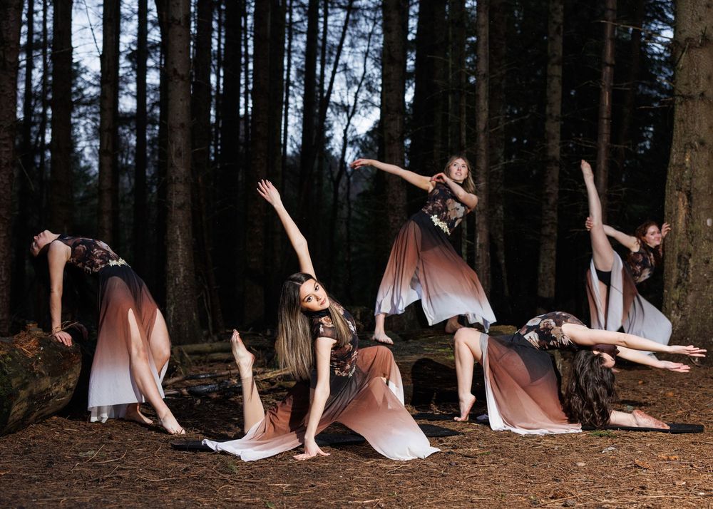 Graceful dancers in flowing ombre gowns perform expressive poses among tall trees in a mystical forest setting.
