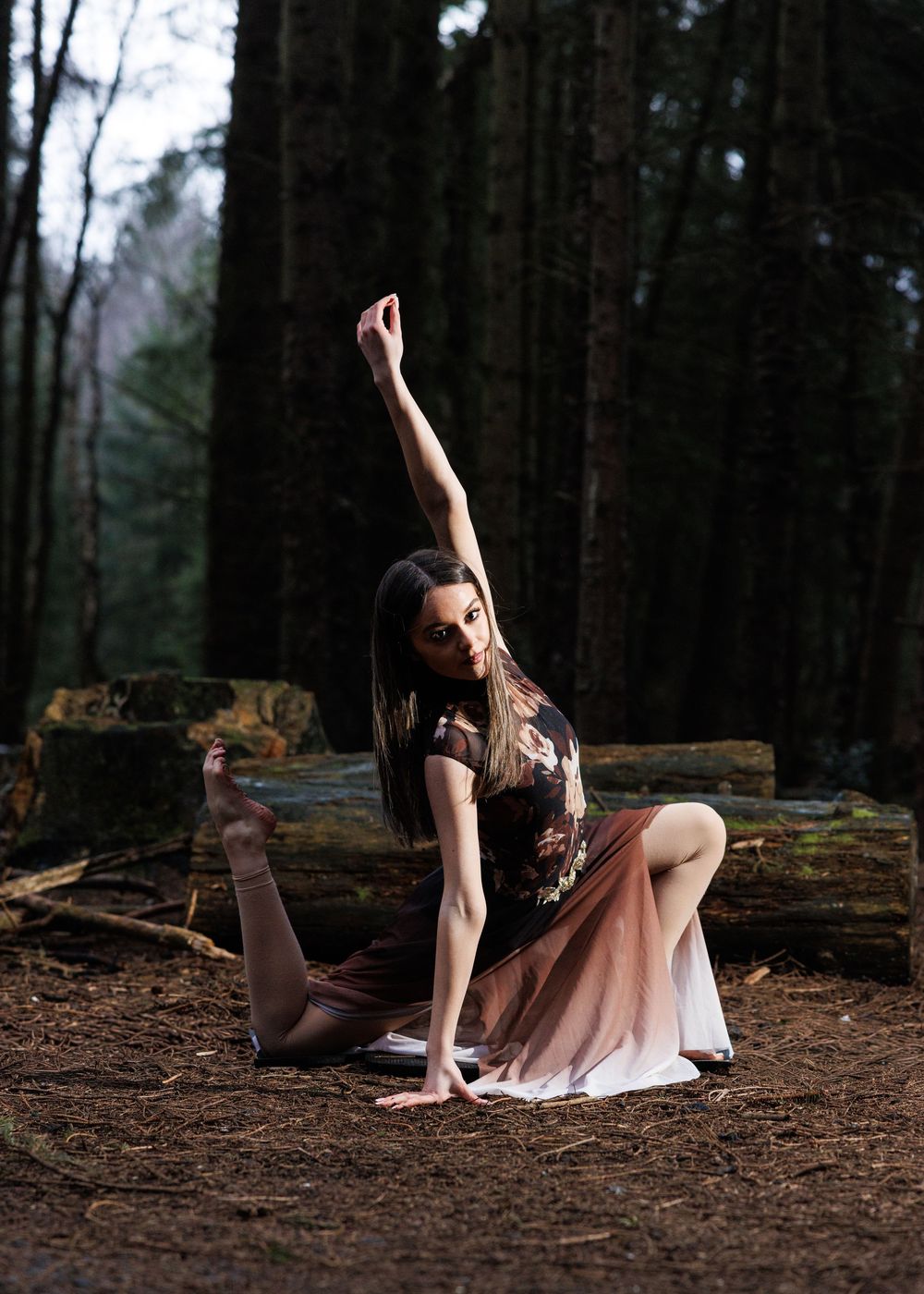 A dancer in a flowing brown ombre dress poses gracefully in a forest, arm raised, seated on the ground.