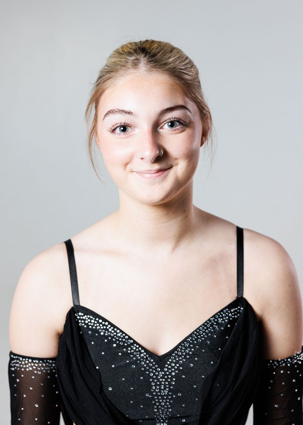 Young woman in black rhinestone dress with sheer gloves, smiling portrait against gray background.