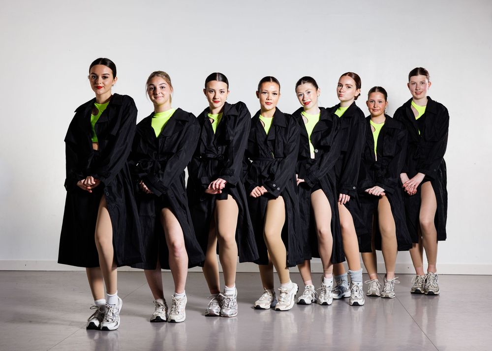 Eight women in matching black coats and neon green tops pose together in a studio, wearing white sneakers.
