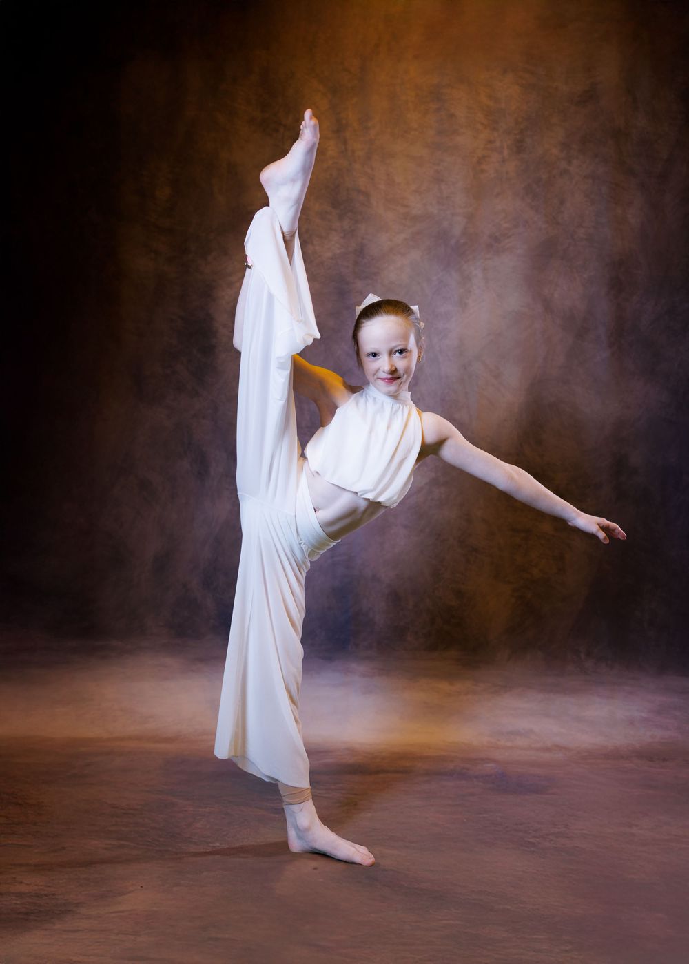 Young dancer in white costume performs a high leg extension pose in a professional studio setting.