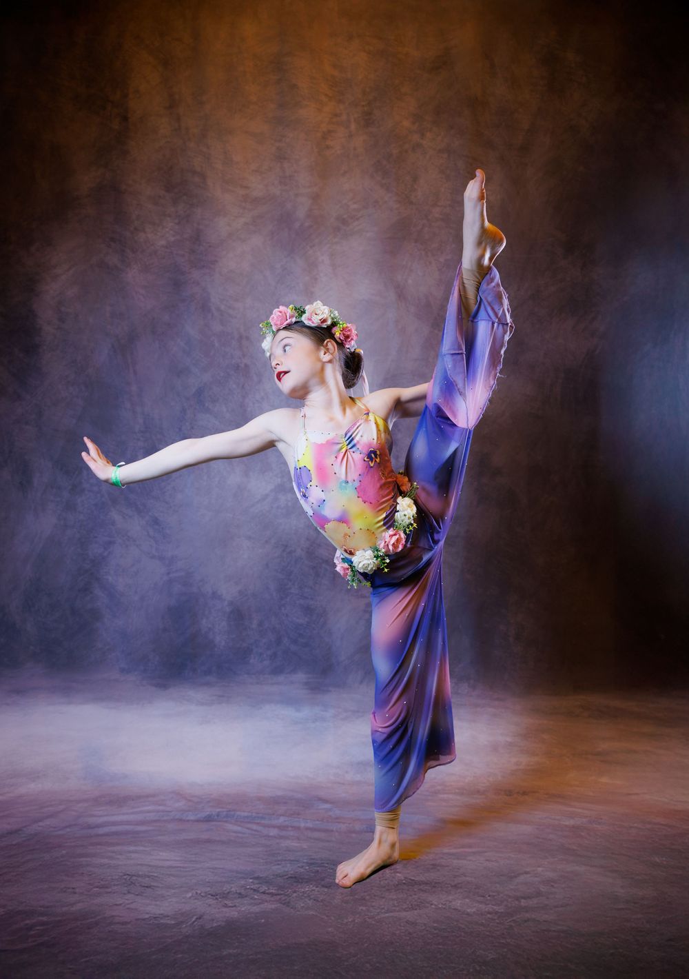 Young dancer in colorful leotard and floral crown performs a high leg extension pose against a moody studio backdrop.