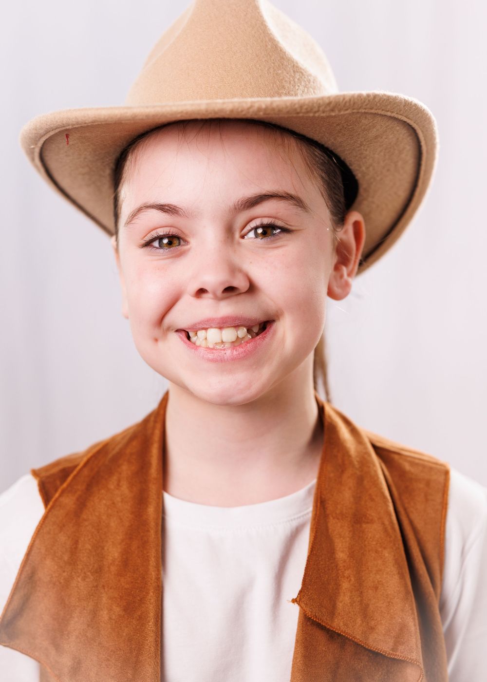 Smiling child wearing a beige cowboy hat and brown suede vest over a white shirt, posing for a portrait.