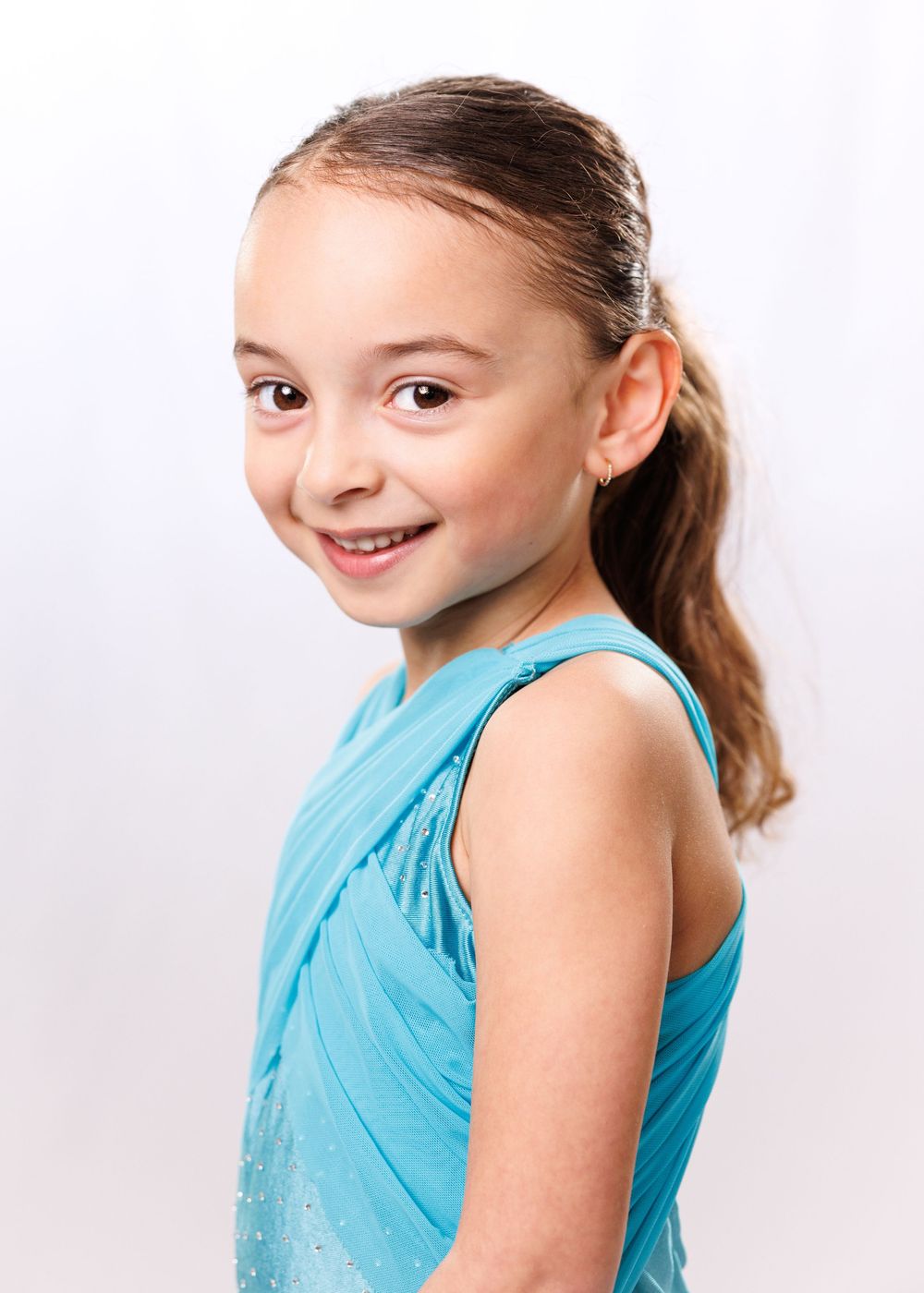 Young girl with ponytail smiling over shoulder, wearing turquoise sleeveless top, studio portrait photo.