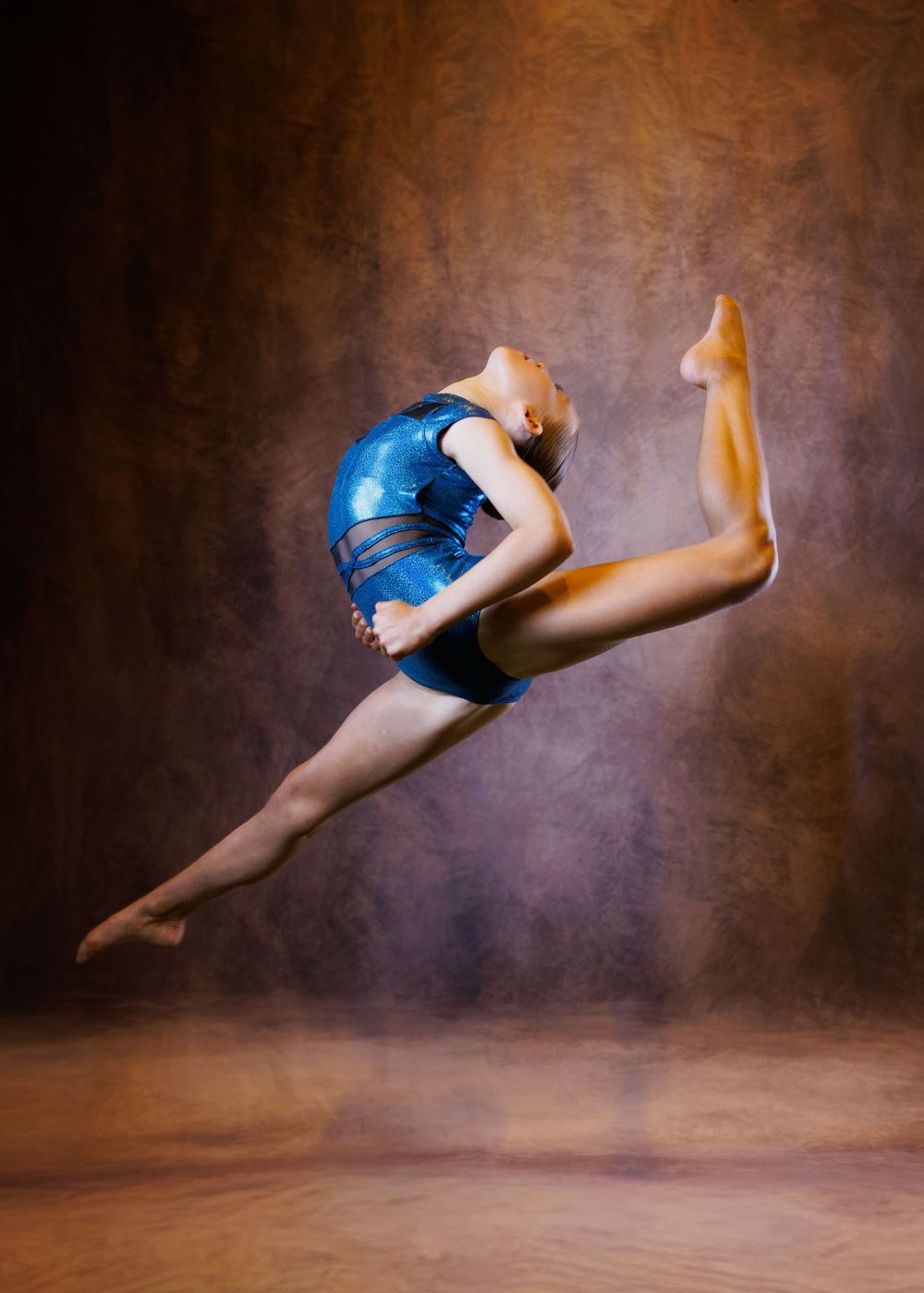 Gymnast in blue leotard performs a stunning mid-air leap with a backbend against a warm brown studio backdrop.